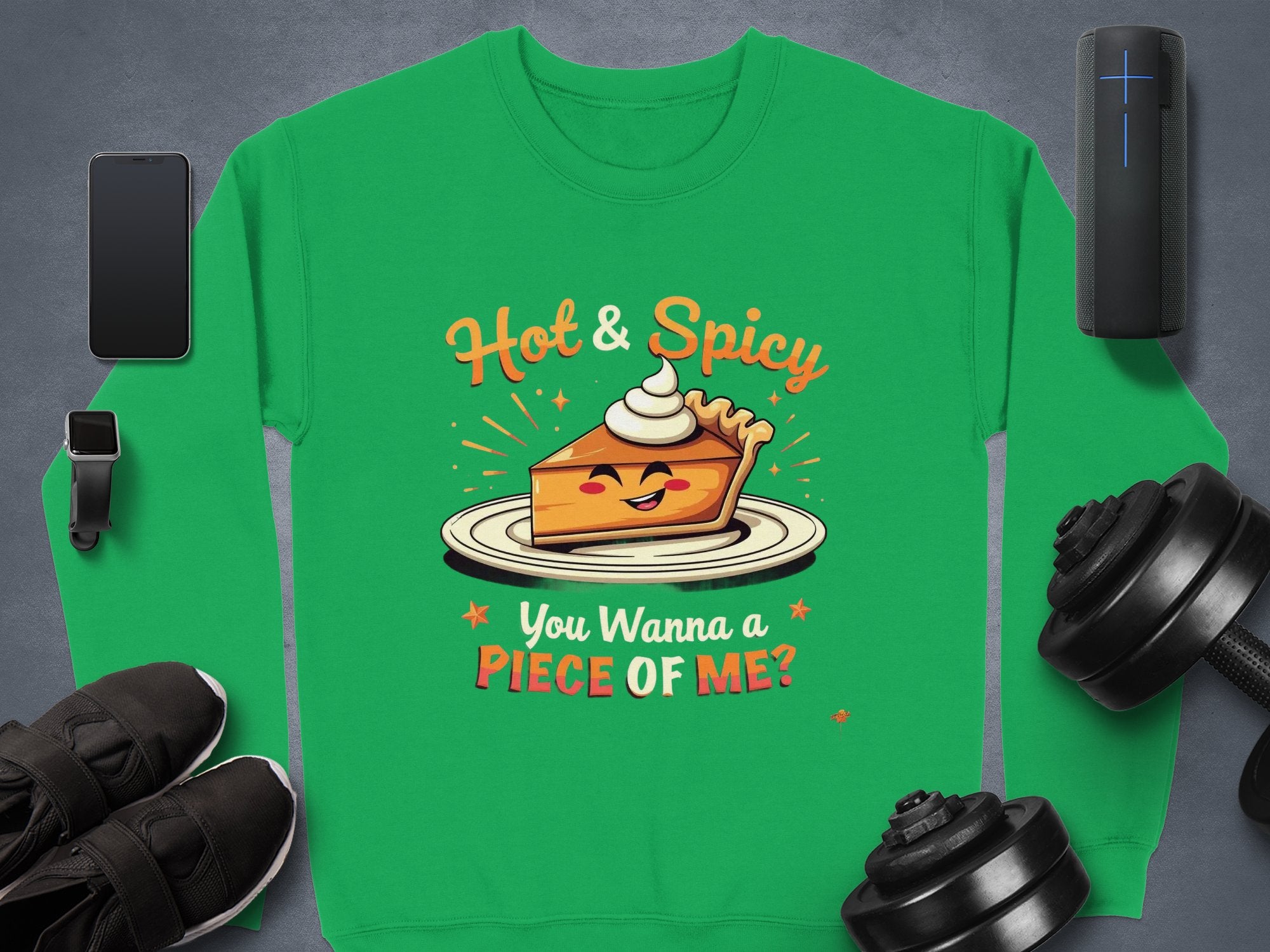 The Garment Graphics pie graphic sweatshirt in vibrant green features a playful illustration of a smiling pie slice with cream, alongside the phrases Hot & Spicy and You Wanna a Piece of Me? Its accompanied by essentials like a smartphone, smartwatch, black shoes, weights, and a speaker.
