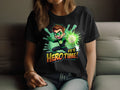 A person lounges on a couch in a Garment Graphics black T-shirt with a green superhero design featuring a cartoon character in a heroic pose, vibrant energy surrounding them, and the phrase Its Hero Time! below, enhancing their colorful wardrobe.