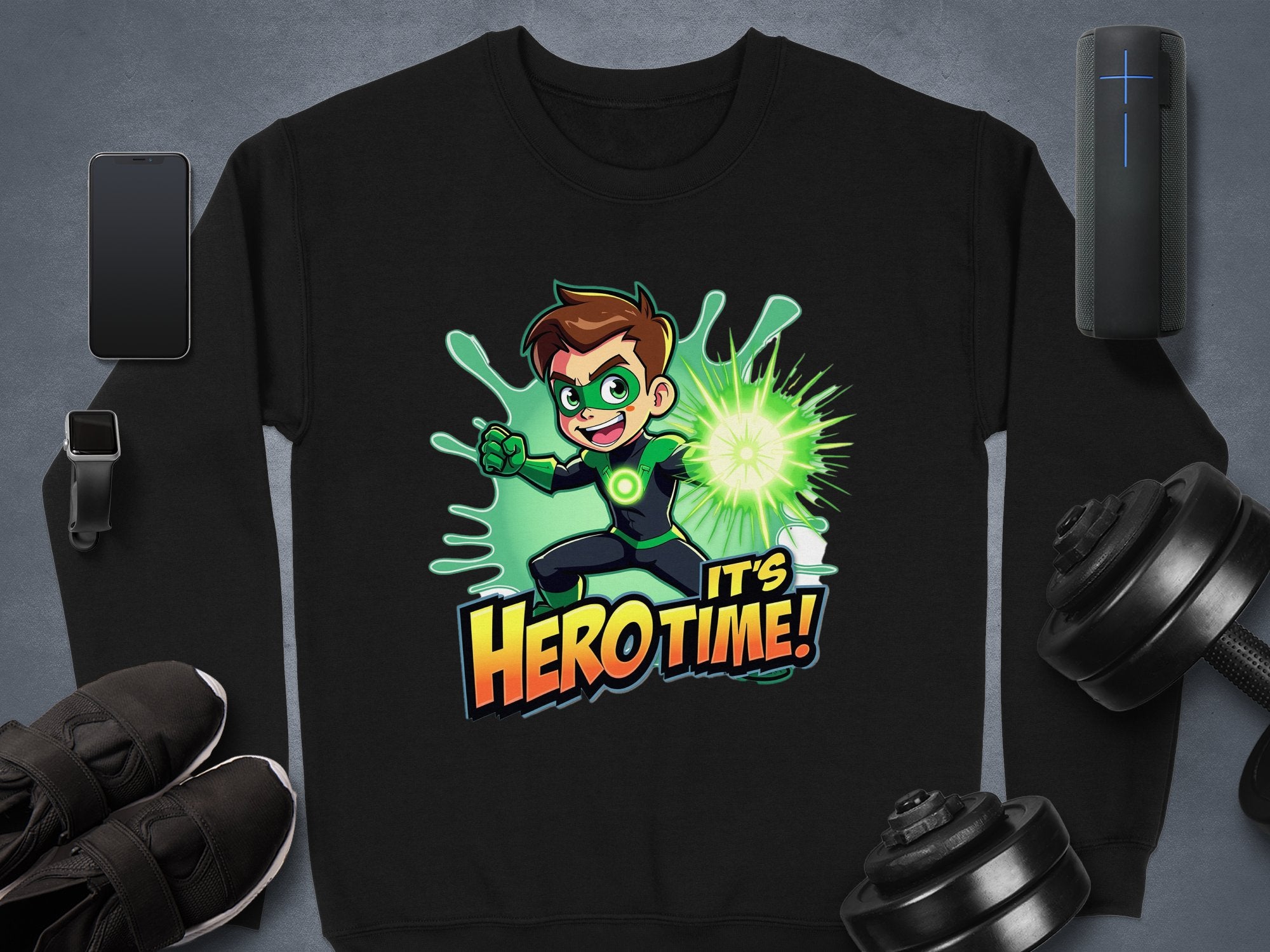 A Garment Graphics black sweatshirt features an animated character with spiky hair, holding a glowing green energy ball amidst a splash effect. Bold Its Herotime! text adds flair for adventure-loving youth. Displayed on gray alongside a watch and weights.