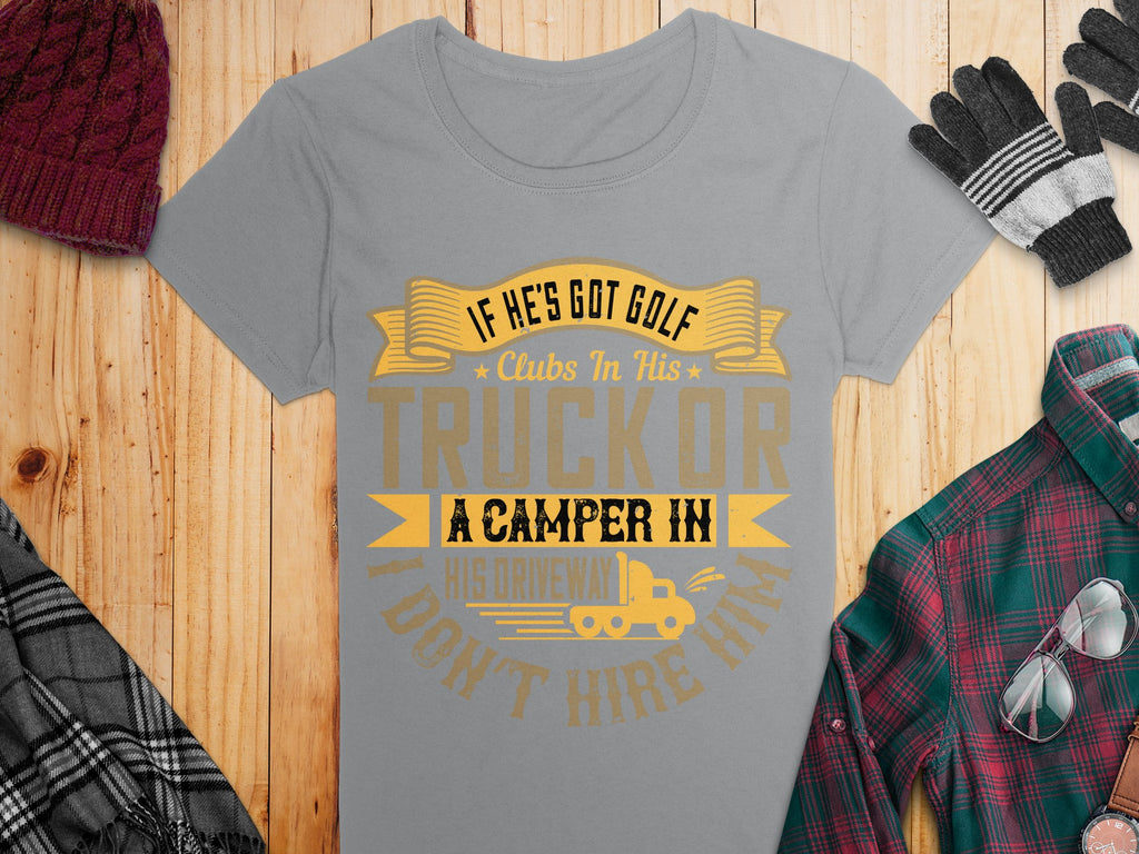Garment Graphics gray graphic tee features the cheeky text If hes got golf clubs in his truck or a camper in his driveway, I dont hire him in gold and black. Nearby on a wooden surface are a red knit hat, black and gray gloves, and a green and red plaid shirt.