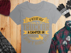 Garment Graphics gray graphic tee features the cheeky text If hes got golf clubs in his truck or a camper in his driveway, I dont hire him in gold and black. Nearby on a wooden surface are a red knit hat, black and gray gloves, and a green and red plaid shirt.