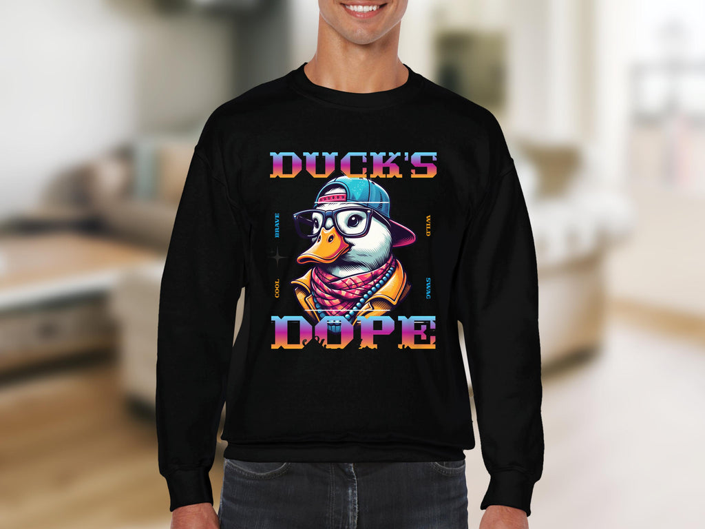 A person is wearing a Garment Graphics black medium-heavy sweatshirt with colorful DUCKS DOPE text, featuring a cartoon duck in a hat, sunglasses, and scarf. Its available in unisex sizing and set against a blurred interior background.