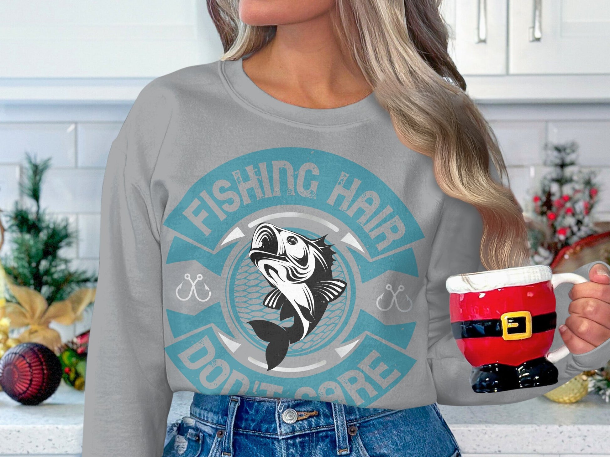 A woman with long hair wears a Garment Graphics fishing sweatshirt featuring Fishing Hair Dont Care and a playful fish print. She holds a Santa-themed mug in her festive kitchen, showcasing her true fishing enthusiasm during the holiday season.