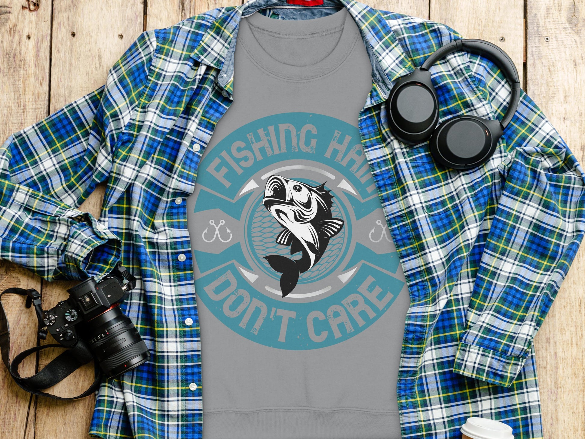 A flat lay features a Garment Graphics gray fishing sweatshirt with the print Fishing Hard Dont Care, tucked under a blue plaid shirt. Surrounding it on the wooden surface are fishing essentials: a camera, headphones, a white thermos, and a smartphone.