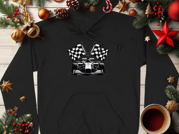 A black Garment Graphics hoodie with a racing theme, featuring checkered flags and a race car graphic, is displayed on a wooden surface. Fir branches, pine cones, cinnamon sticks, ornaments, and a steaming cup of coffee surround it for a festive touch.