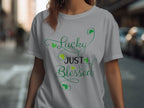 A person is wearing a classic fit light gray t-shirt from Royal Mail - 24 Tracked, featuring Lucky Just Blessed in green cursive with small shamrock graphics. They stand on a street with blurred buildings and cars in the background, enhancing the charm of their graphic t-shirt.