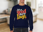 A person wears a medium-heavy navy blue sweatshirt by Garment Graphics, featuring Its a Good Day to Laugh in bold, colorful letters. The blurred neutral-toned indoor background enhances the cozy appeal of this graphic printed sweatshirt.