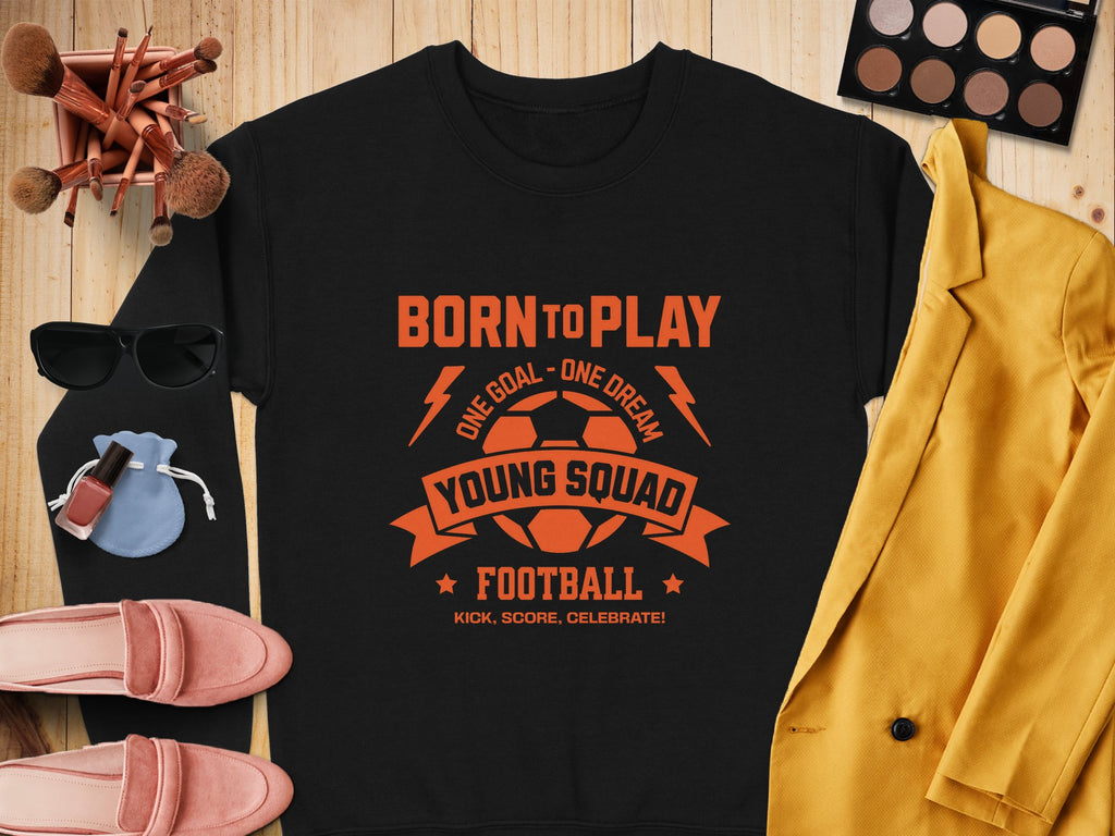 Garment Graphics black unisex t-shirt features an orange football design that says Born to Play, One Goal - One Dream, Young Squad Football, Kick, Score, Celebrate! It’s styled with makeup brushes, a palette, sunglasses, a blue pouch, perfume, shoes, and a yellow jacket.