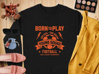 Garment Graphics black unisex t-shirt features an orange football design that says Born to Play, One Goal - One Dream, Young Squad Football, Kick, Score, Celebrate! It’s styled with makeup brushes, a palette, sunglasses, a blue pouch, perfume, shoes, and a yellow jacket.