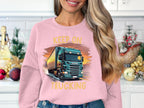 A long-haired individual stands in a kitchen, showcasing their love for trucks by wearing a Garment Graphics sweatshirt that reads Keep on Trucking. Festive ornaments and a small Christmas tree add to the cheerful backdrop.