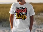 Wearing a Garment Graphics Its a Good Day to Laugh T-shirt, a person casually stands with hands in pockets. The shirts vibrant colors amplify the relaxed, outdoor vibe against sandy beach and grassy dunes.