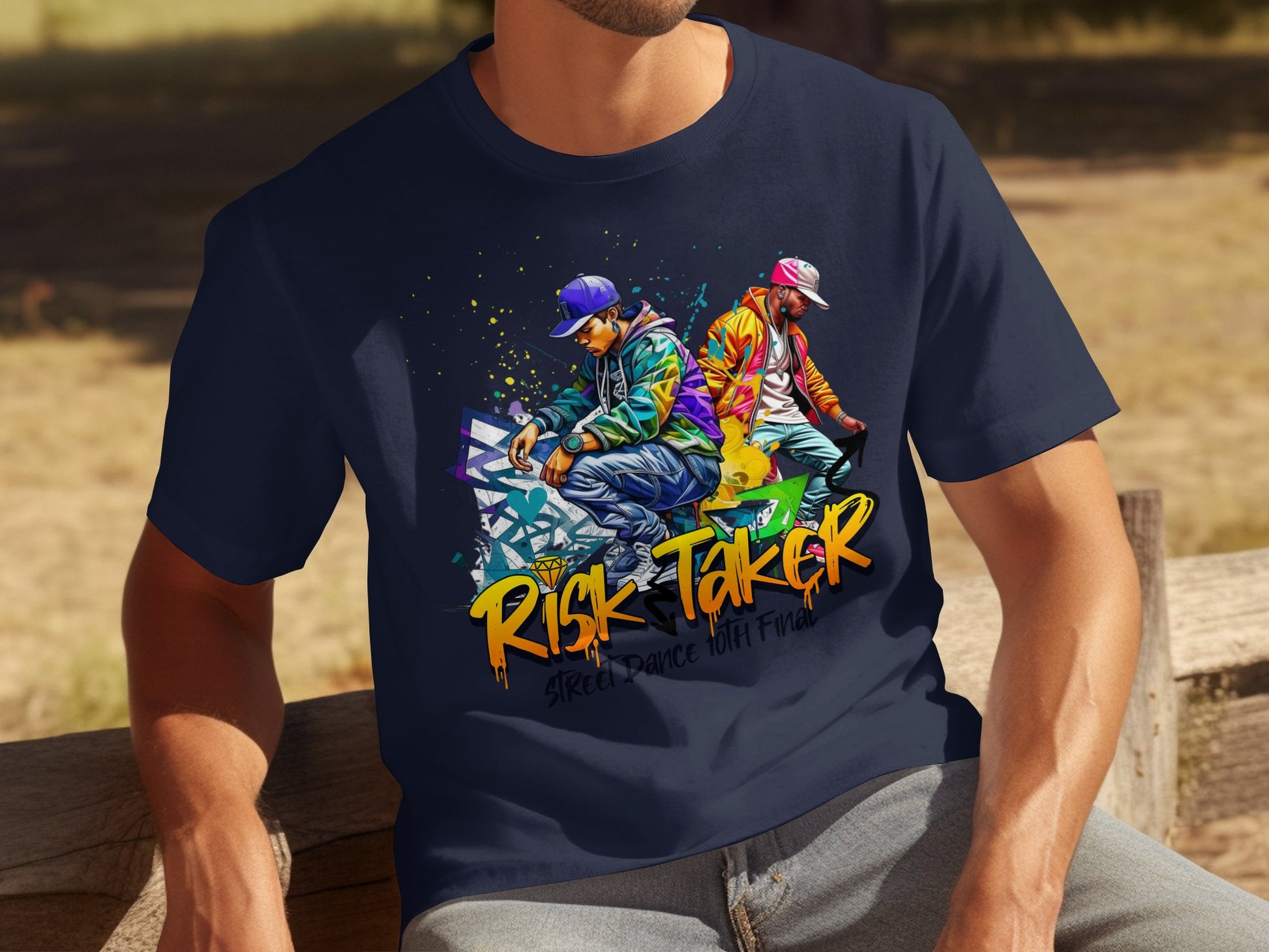 A man wears a Garment Graphics dark blue t-shirt adorned with lively graffiti art of two dancers and the phrase Risk Taker, Street Dance with Friends. Set against a grassy backdrop, this piece honors street dance culture.