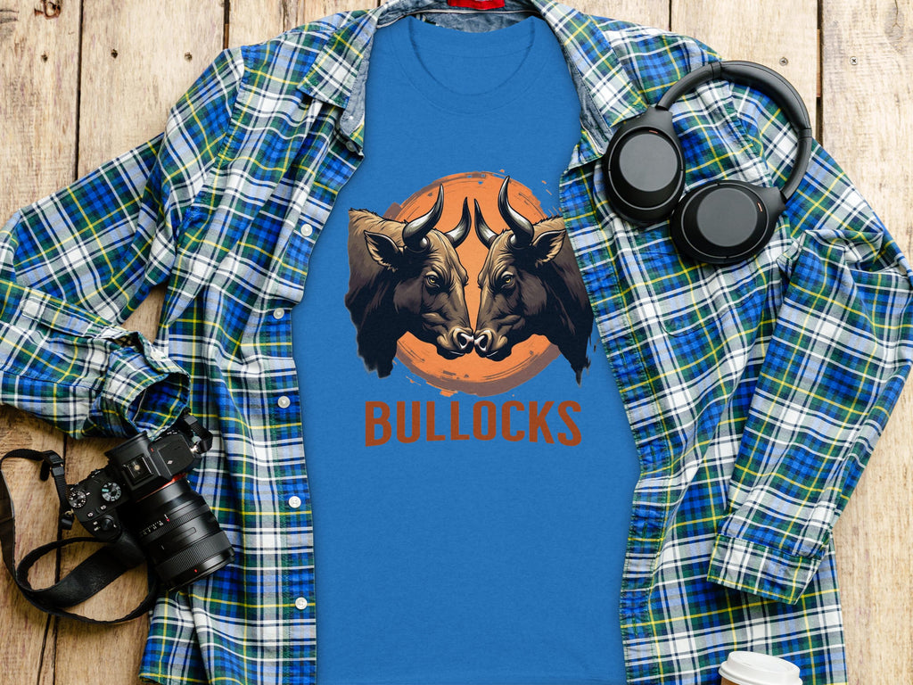 A blue Garment Graphics t-shirt with a double bull head design and BULLOCKS is displayed on a plaid shirt on wood. Nearby lie headphones, a camera, and a takeaway coffee cup. This classic fit tee is ideal for family sizing.