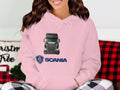 A woman in a pink Garment Graphics Scania Truck Hoodie featuring a truck print smiles. The scene is cozy with a red and black plaid blanket ideal for truck enthusiasts, alongside part of a Christmas tree.