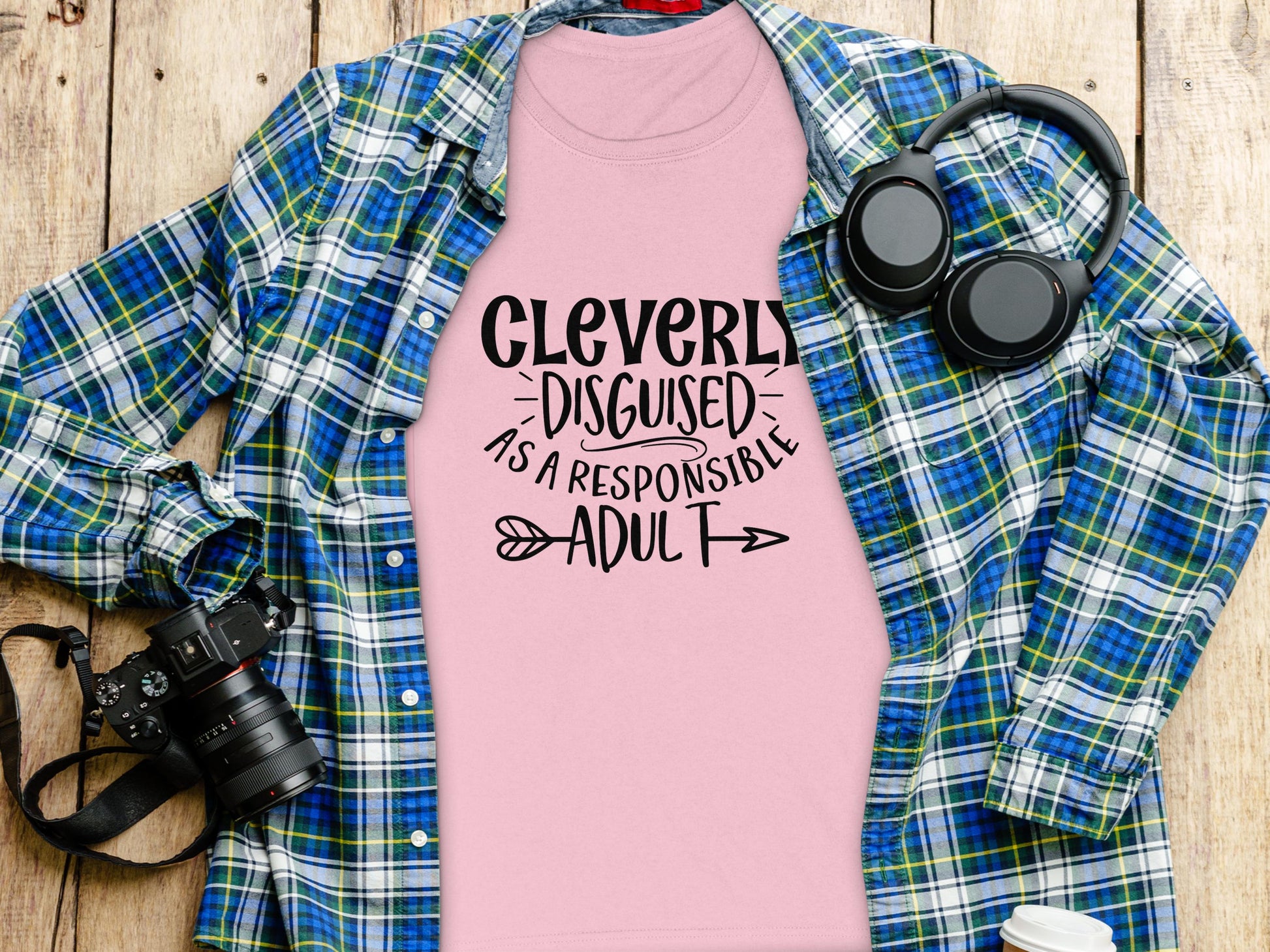 A semi-fitted pink T-shirt by Garment Graphics, featuring Cleverly Disguised as a Responsible Adult, lies on wood. Nearby, a blue and green plaid shirt complements a black camera and slick black headphones, epitomizing effortless style.