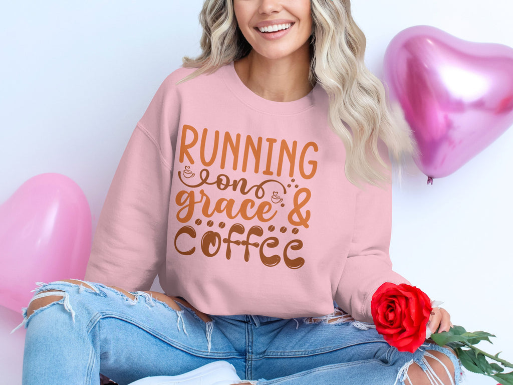 A person sits cross-legged in a Garment Graphics pink graphic sweatshirt with Running on Grace & Coffee in unisex sizing. Holding a red rose, theyre surrounded by pink heart-shaped balloons, their long wavy hair cascading over ripped jeans.