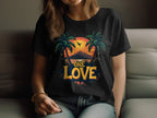 A woman relaxes on a couch in jeans and a Garment Graphics black t-shirt showcasing vibrant palm trees, a tropical sunset, and a plane silhouette. One Love is boldly printed beneath, giving her a casual beach-ready vibe.