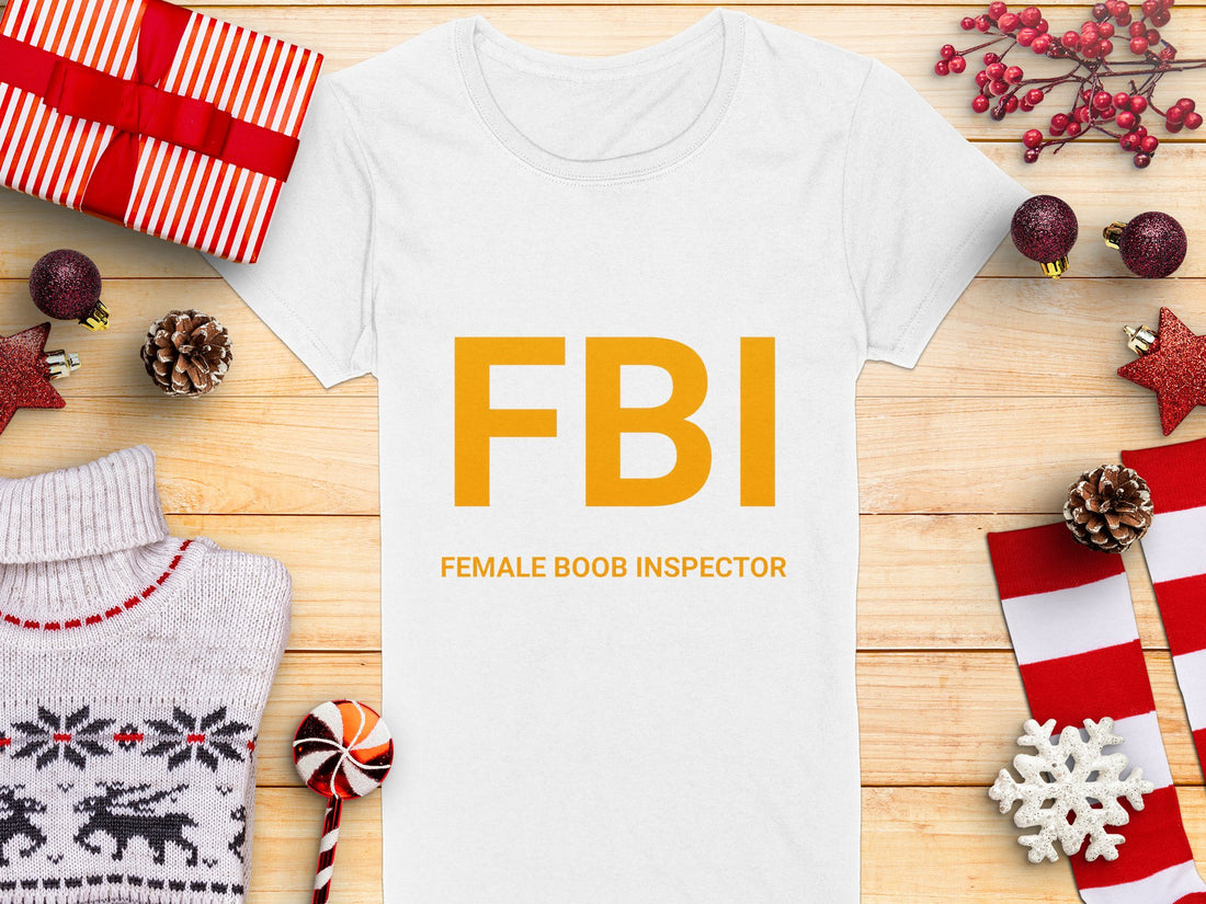 A white Garment Graphics T-shirt features yellow FBI Female Boob Inspector text, lying on wood. Its surrounded by holiday items: gifts, a deer-patterned sweater, pine cones, red ornaments, and a candy cane. Ideal as a gag gift and available in womens sizes.