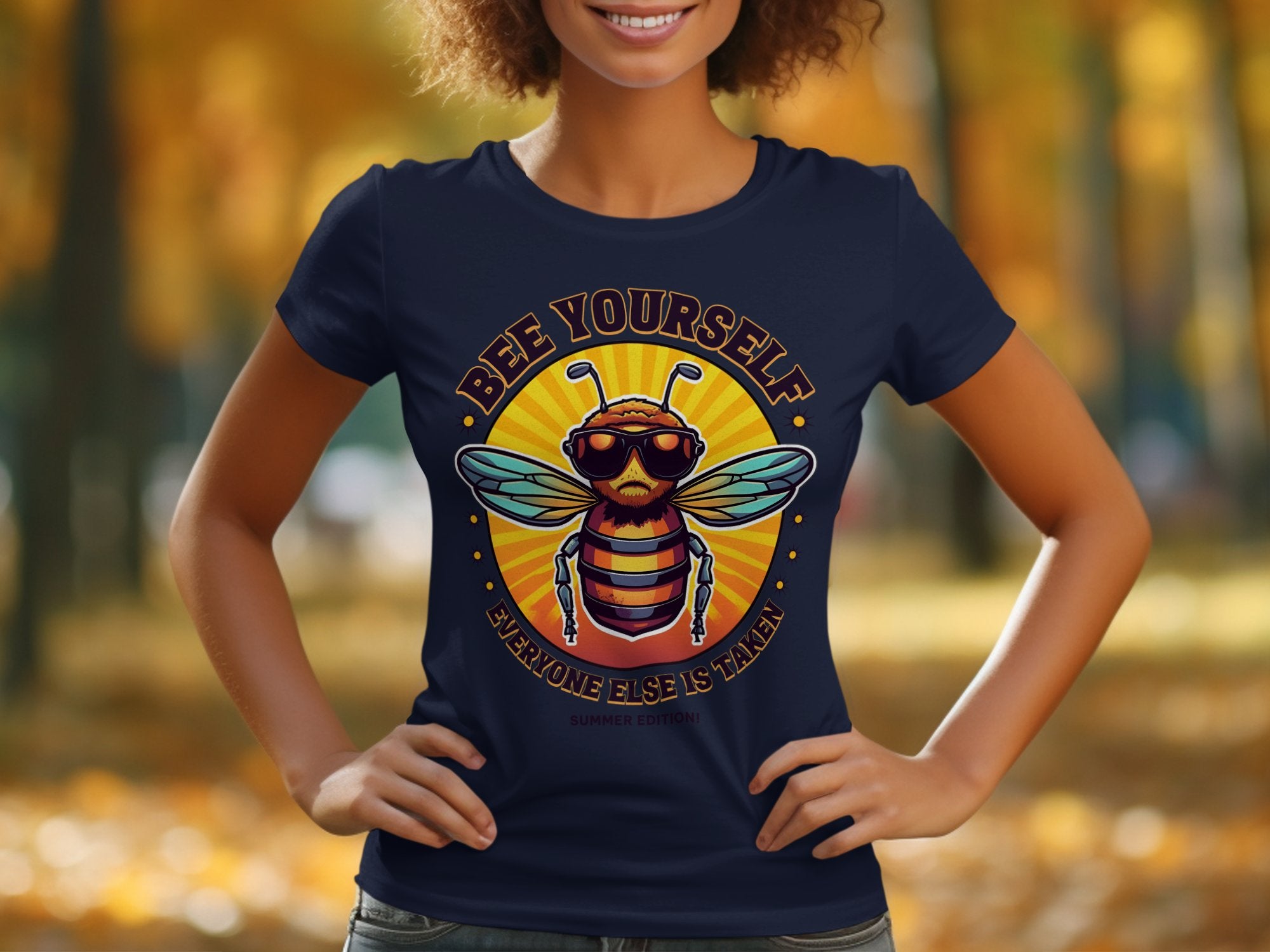 Outdoors amidst blurred autumn foliage, a person wears a Garment Graphics Bee Yourself Navy Womens T-Shirt featuring a bee graphic and the text Bee Yourself, Everyone Else is Taken, Summer Edition.