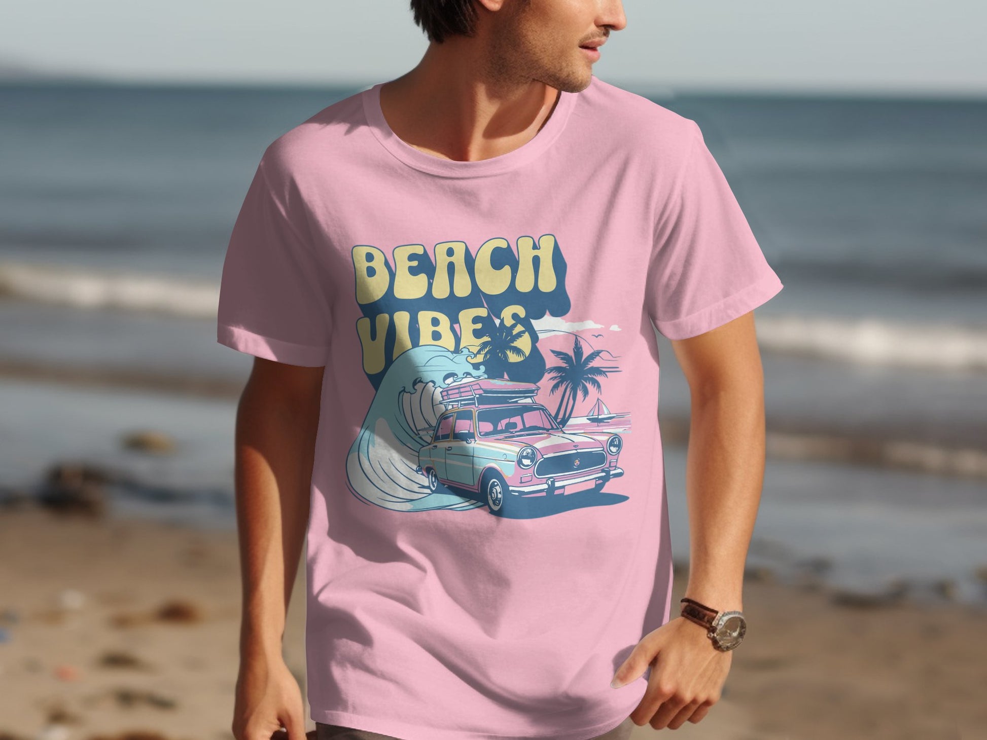 A person on the beach is wearing a Garment Graphics pink t-shirt featuring a classic car, palm trees, waves, and bold Beach Vibes lettering, perfectly complementing the ocean and sand backdrop.
