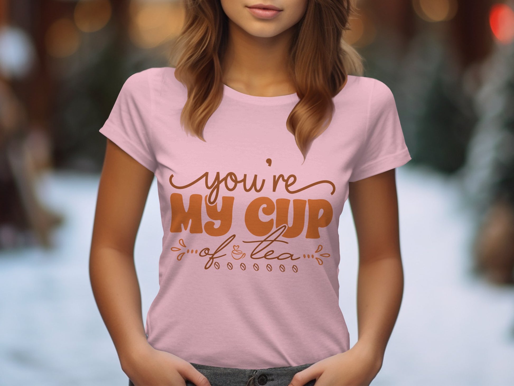 A woman wears a Garment Graphics 100% cotton graphic T-shirt featuring youre my cup of tea in brown. She stands outside against a backdrop of blurred trees and snow, with her hands cozily tucked into her pockets.