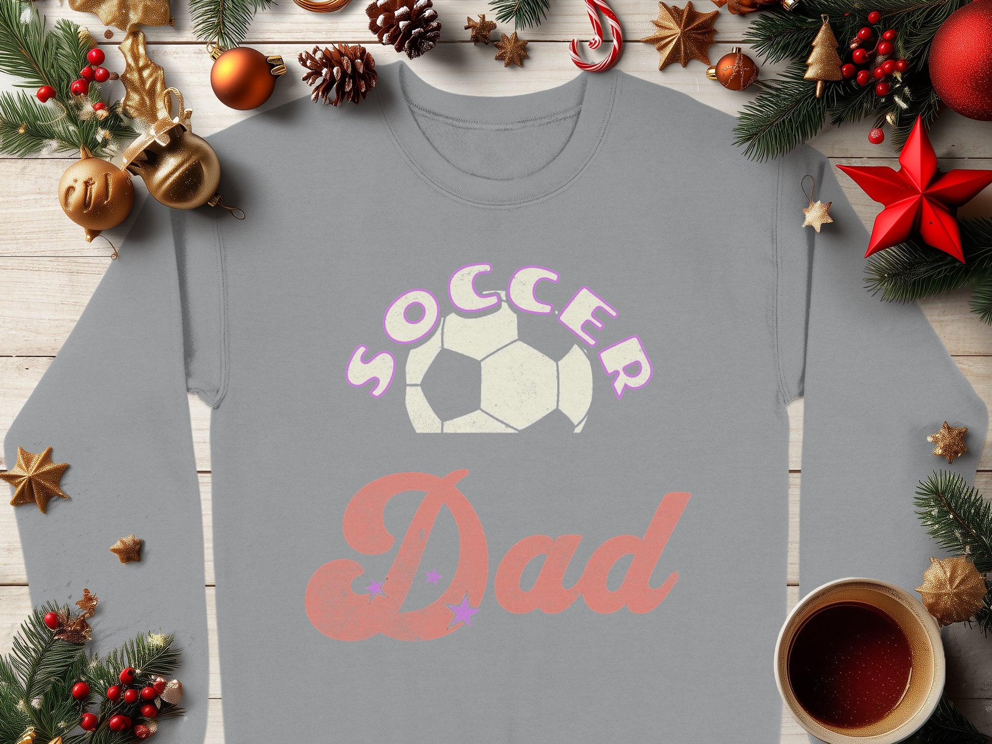 A Garment Graphics Soccer Dad sweatshirt with a soccer ball graphic lies on wood, adorned with pine branches, red and gold ornaments, festive pinecones, a candy cane, and a steaming coffee cup—perfect for all in unisex sizing.