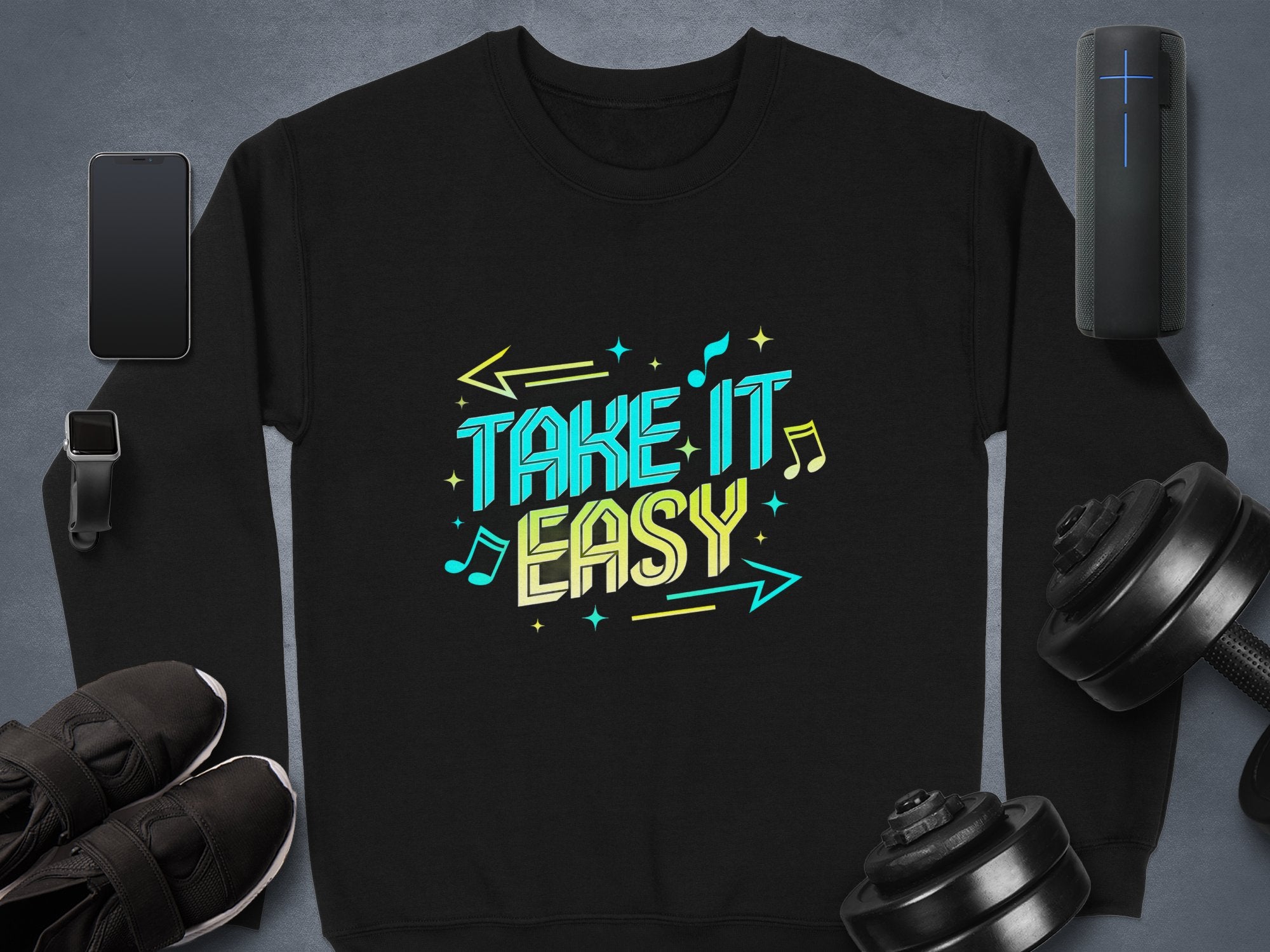Garment Graphics black sweatshirt features Take It Easy in bold, colorful letters—an epitome of graphic design. On a gray surface, its surrounded by a smartphone, smartwatch, wireless speaker, black shoes, and dumbbells with musical notes and arrows enhancing the text.