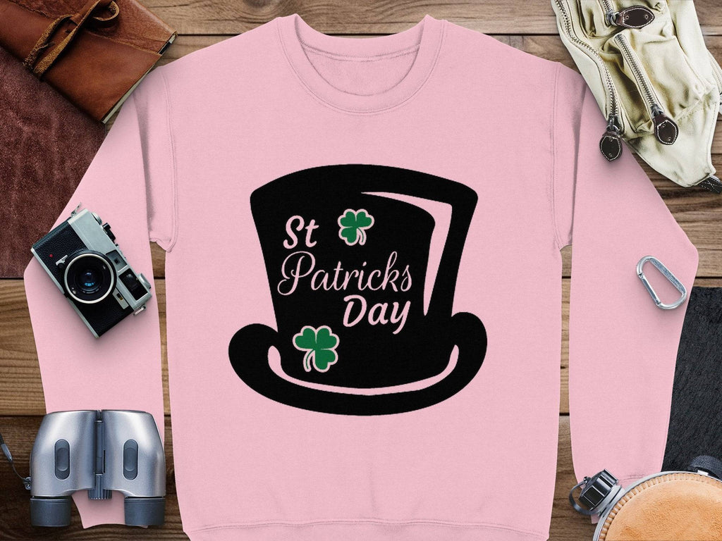 A Garment Graphics pink sweatshirt featuring a black graphic of a top hat and shamrocks with St Patricks Day text captures the festive spirit. Its styled on a wooden surface with a vintage camera, binoculars, brown leather wallet, and climbing gear.