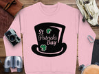 A Garment Graphics pink sweatshirt featuring a black graphic of a top hat and shamrocks with St Patricks Day text captures the festive spirit. Its styled on a wooden surface with a vintage camera, binoculars, brown leather wallet, and climbing gear.