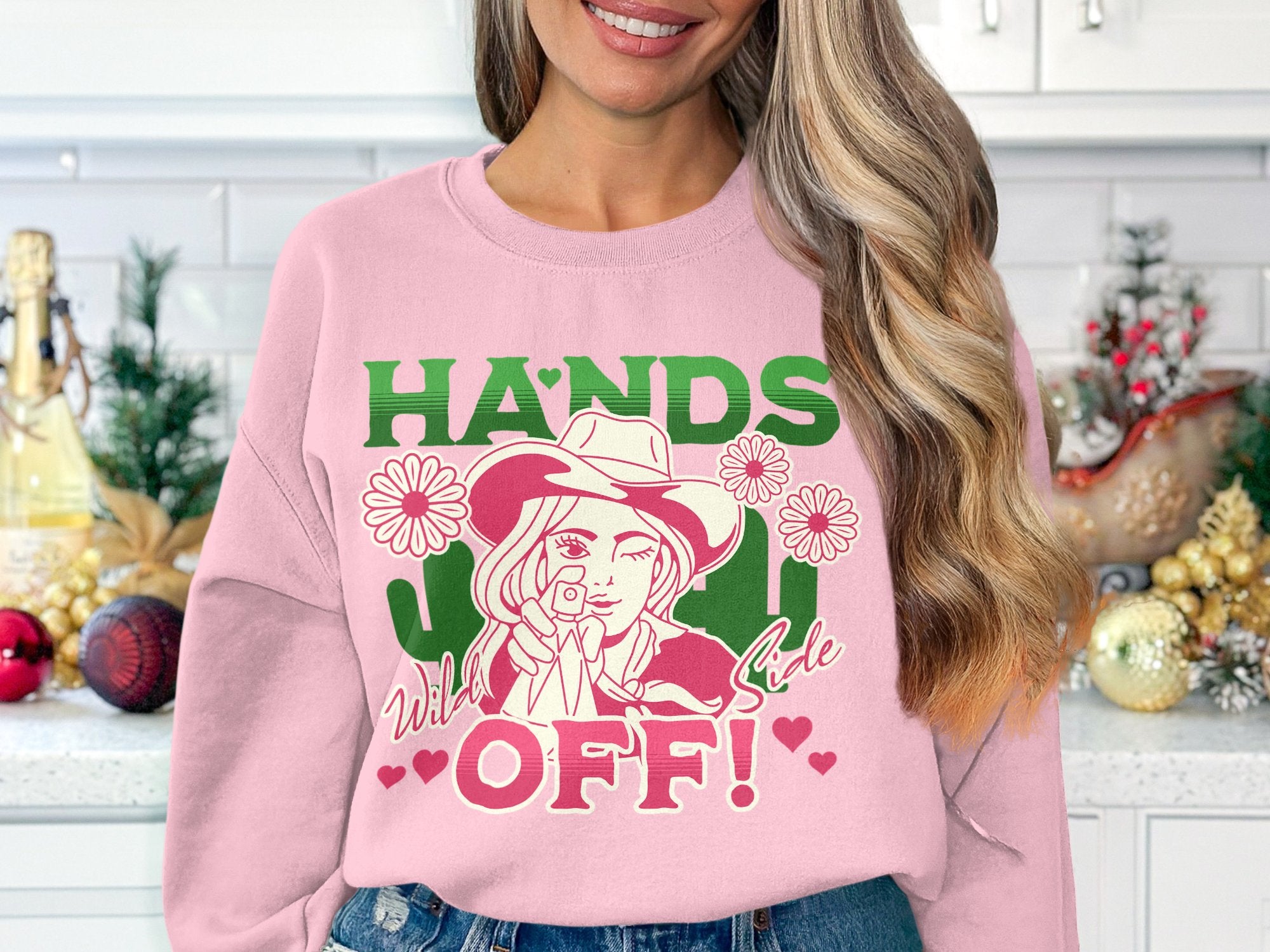 A woman in a kitchen wears a pink Garment Graphics Hands Off Wild Side Sweatshirt with HANDS OFF! boldly displayed. The design shows a woman in a cowboy hat amid flowers, and its comfy loose fit beautifully complements the festive holiday decor behind her on the countertop.