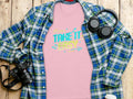 A pink T-shirt from Garment Graphics featuring Take It Easy in vibrant designs is displayed beneath a blue plaid shirt with black headphones, a camera, and a lidded cup nearby, all set on a wooden surface.