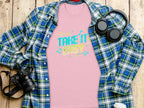 A pink T-shirt from Garment Graphics featuring Take It Easy in vibrant designs is displayed beneath a blue plaid shirt with black headphones, a camera, and a lidded cup nearby, all set on a wooden surface.