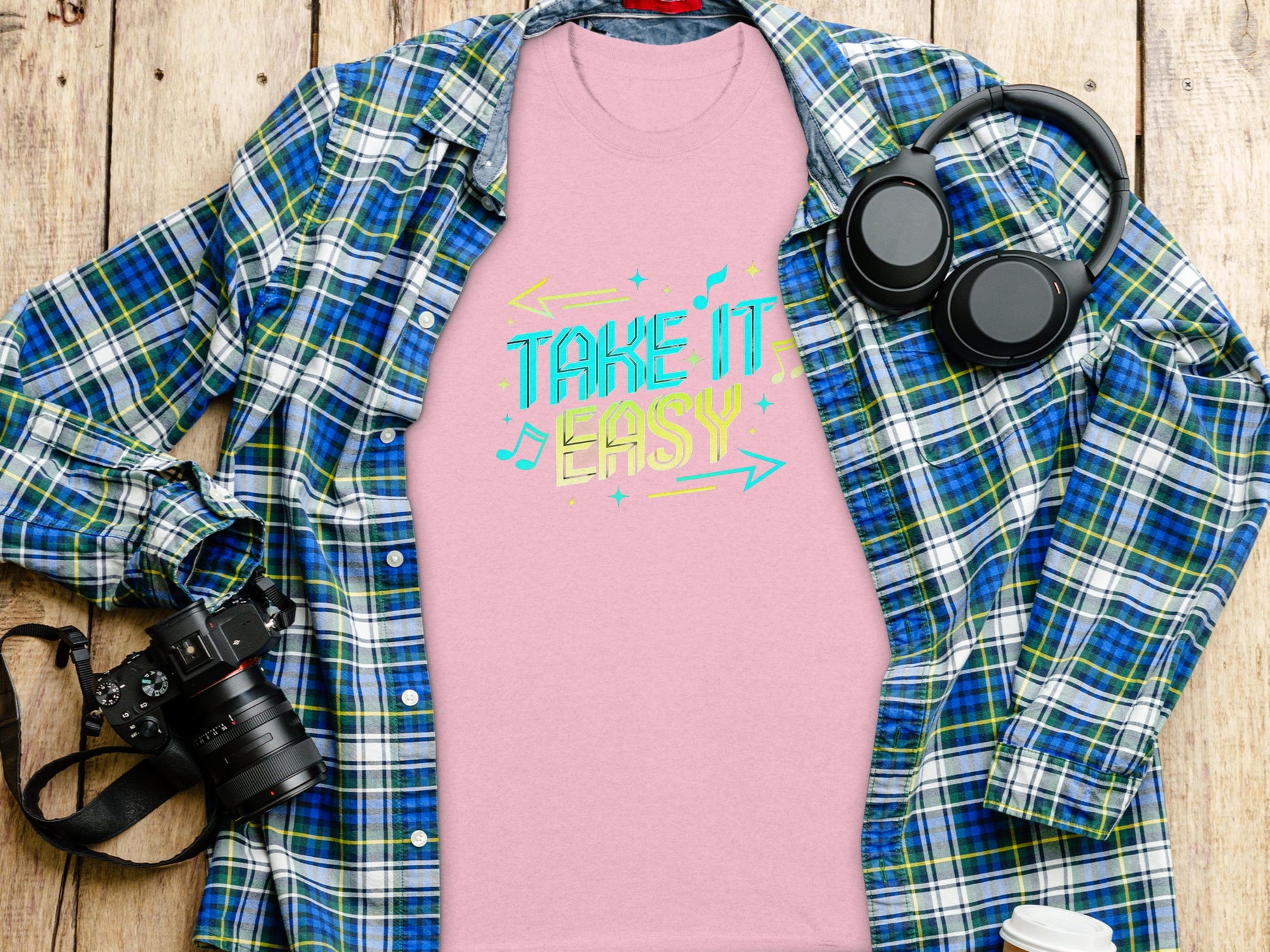 A pink T-shirt from Garment Graphics featuring Take It Easy in vibrant designs is displayed beneath a blue plaid shirt with black headphones, a camera, and a lidded cup nearby, all set on a wooden surface.