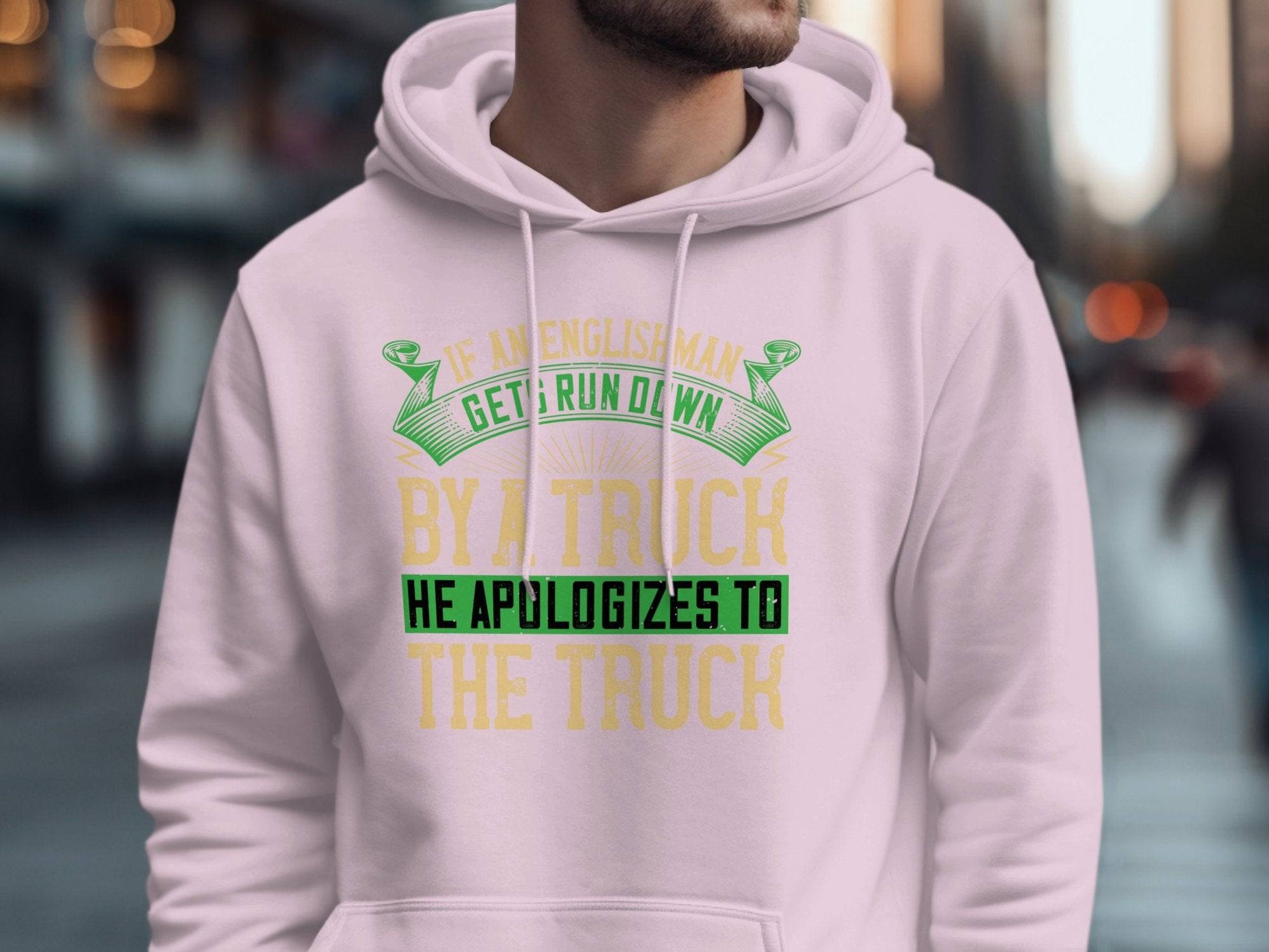 A person on a city street wears a light pink Garment Graphics hoodie featuring the funny quote, If an Englishman gets run down by a truck, he apologizes to the truck, in green, yellow, and black lettering. The humorous design captures English etiquette with its clever twist against a blurred background.