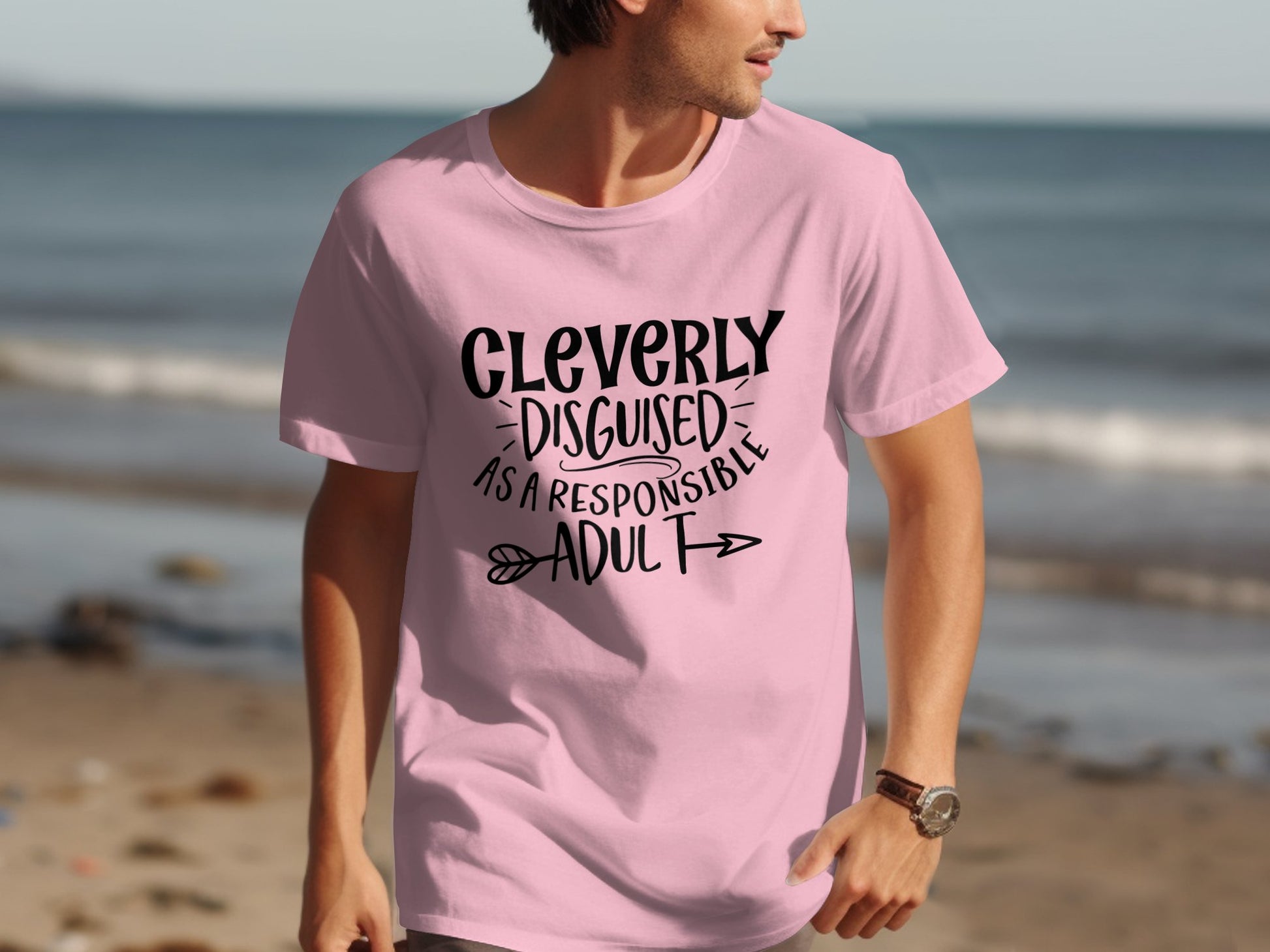 On a casual beach outing, someone strolls along the sand in a Garment Graphics light pink classic fit T-shirt with Cleverly Disguised as a Responsible Adult printed in bold black and decorative arrows, against the picturesque ocean backdrop.