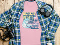 A stylish Garment Graphics pink graphic t-shirt, showcasing Beach Vibes with a classic car, surfing wave, and palm trees, is layered over a plaid shirt. Nearby on the wooden surface: a camera, wireless headphones, and a coffee cup lid—essentials for capturing relaxed moments.