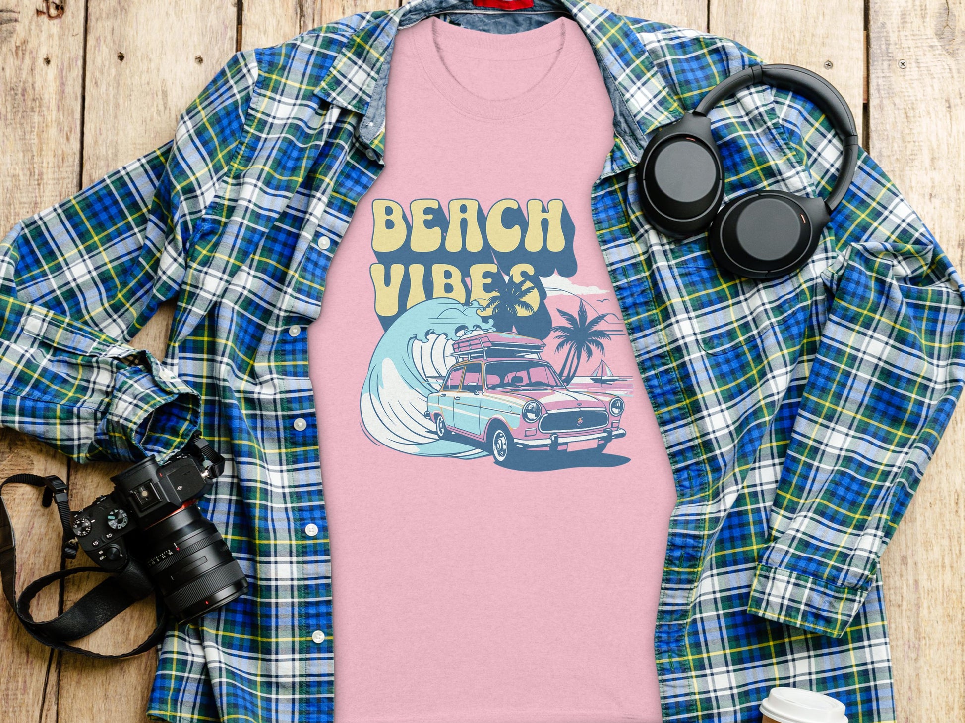 A stylish Garment Graphics pink graphic t-shirt, showcasing Beach Vibes with a classic car, surfing wave, and palm trees, is layered over a plaid shirt. Nearby on the wooden surface: a camera, wireless headphones, and a coffee cup lid—essentials for capturing relaxed moments.