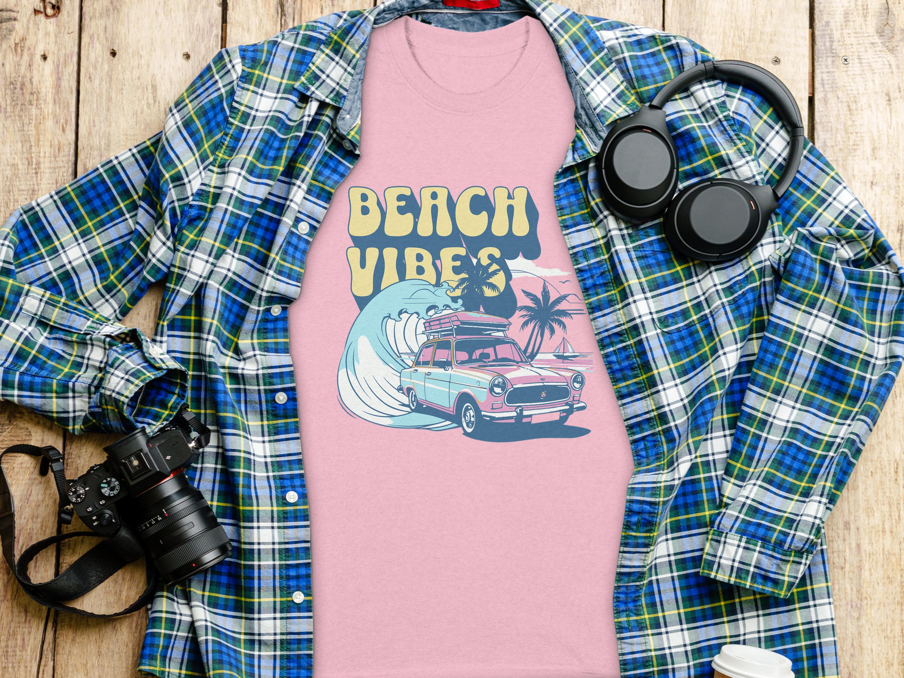 A stylish Garment Graphics pink graphic t-shirt, showcasing Beach Vibes with a classic car, surfing wave, and palm trees, is layered over a plaid shirt. Nearby on the wooden surface: a camera, wireless headphones, and a coffee cup lid—essentials for capturing relaxed moments.