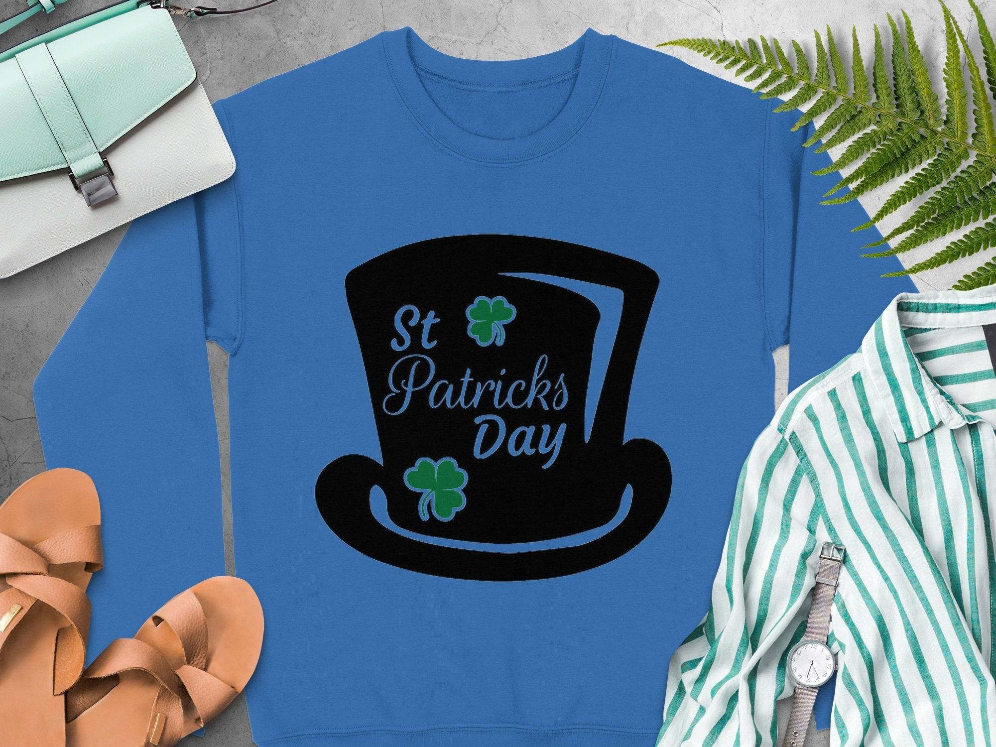 Garment Graphics festive blue sweatshirt with a black top hat, St Patricks Day text, and green shamrocks is styled alongside tan sandals, a mint purse, a striped shirt, a watch, and green ferns on gray.