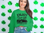 A person with long hair wears a stylish Garment Graphics green sweatshirt featuring a classic car and Enjoy Classic Car, Vintage Ride Community design, set against green shamrocks. They pair it with denim shorts for a trendy look.