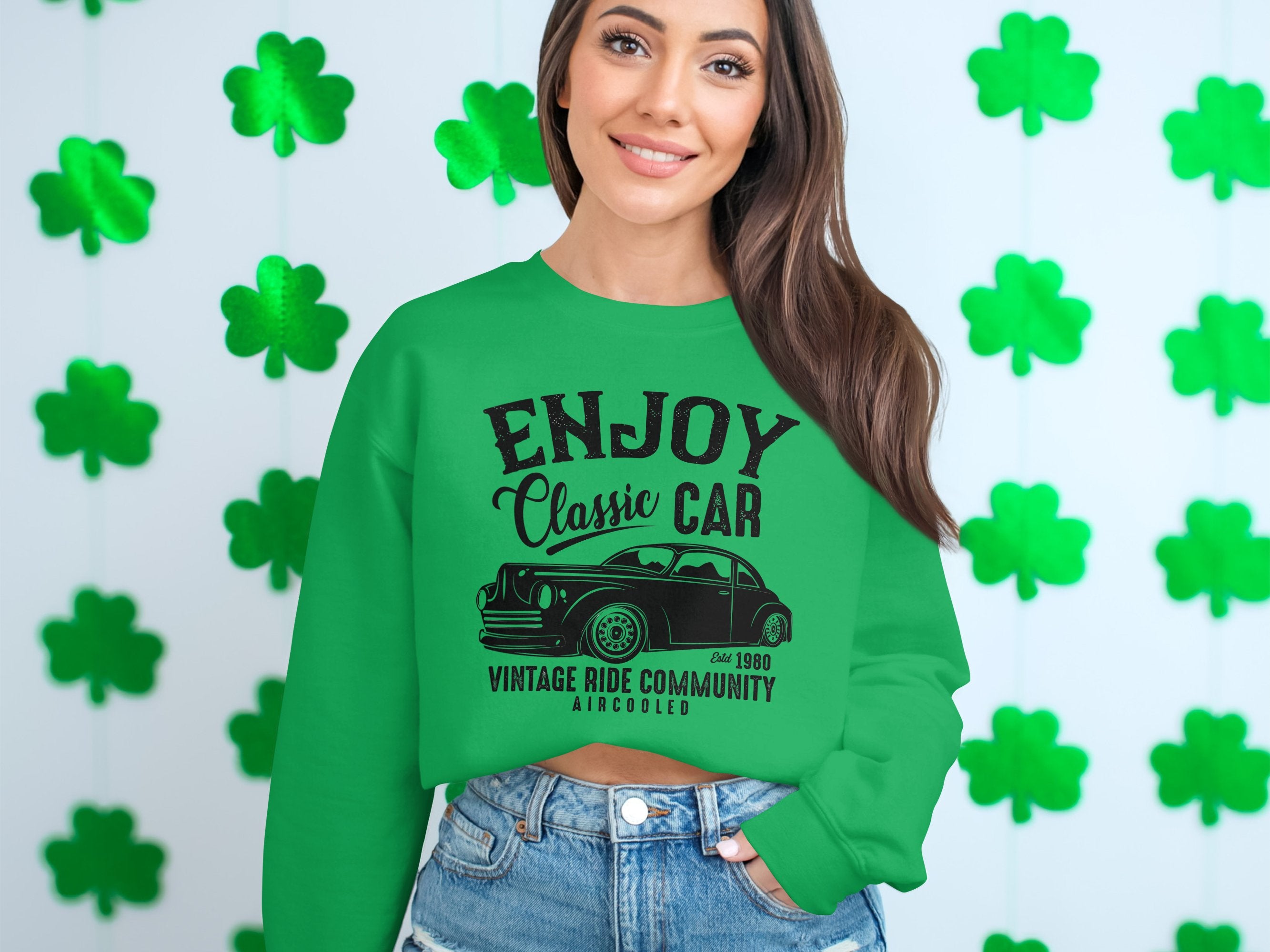 A person with long hair wears a stylish Garment Graphics green sweatshirt featuring a classic car and Enjoy Classic Car, Vintage Ride Community design, set against green shamrocks. They pair it with denim shorts for a trendy look.