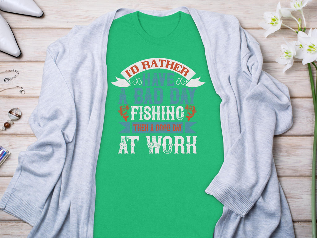 A green Garment Graphics fishing T-shirt with Id rather have a bad day fishing than a good day at work lies on wood, paired with a light gray sweater, flowers, and jewelry—an ideal gift for any angler.