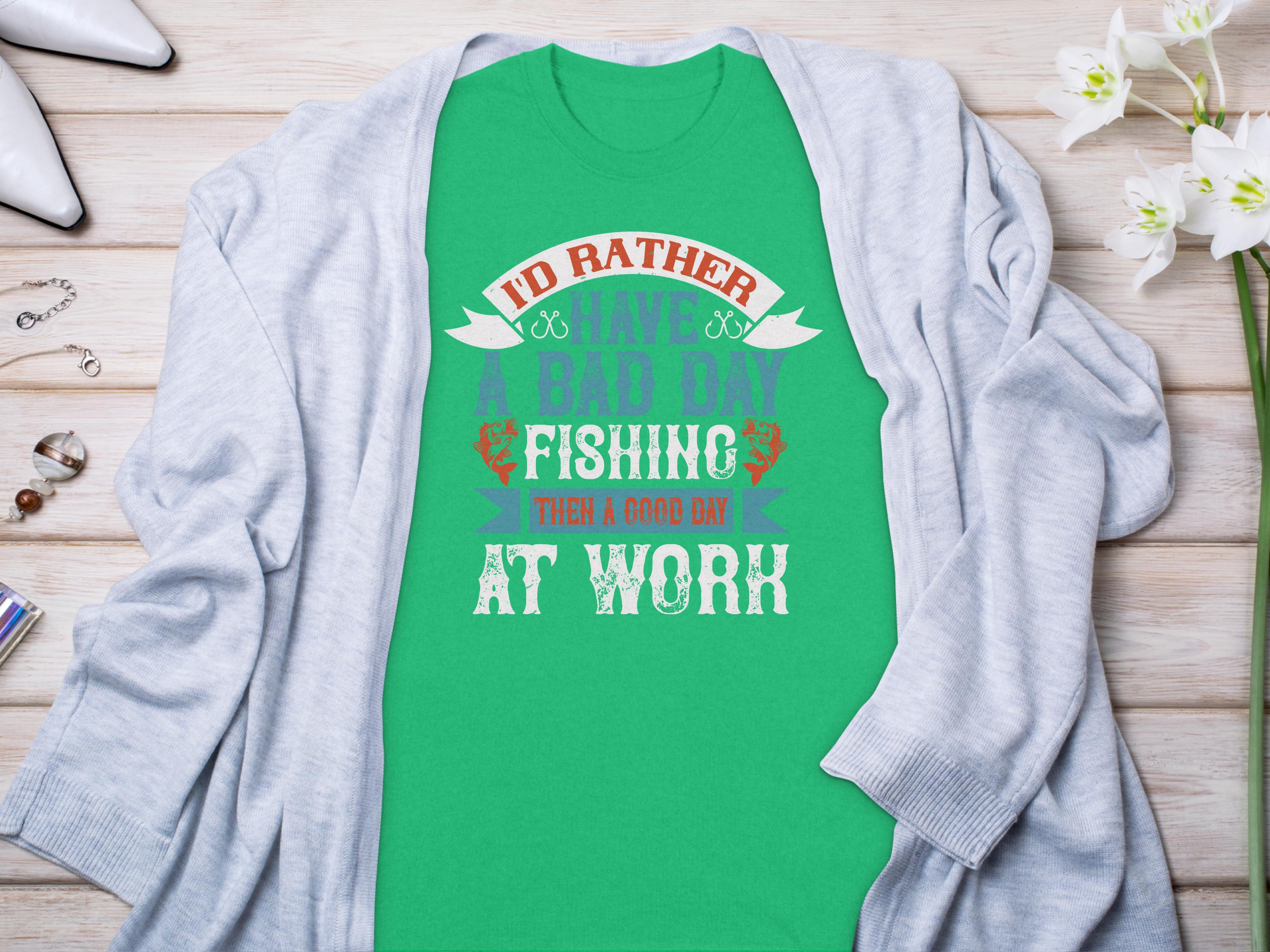 A green Garment Graphics fishing T-shirt with Id rather have a bad day fishing than a good day at work lies on wood, paired with a light gray sweater, flowers, and jewelry—an ideal gift for any angler.