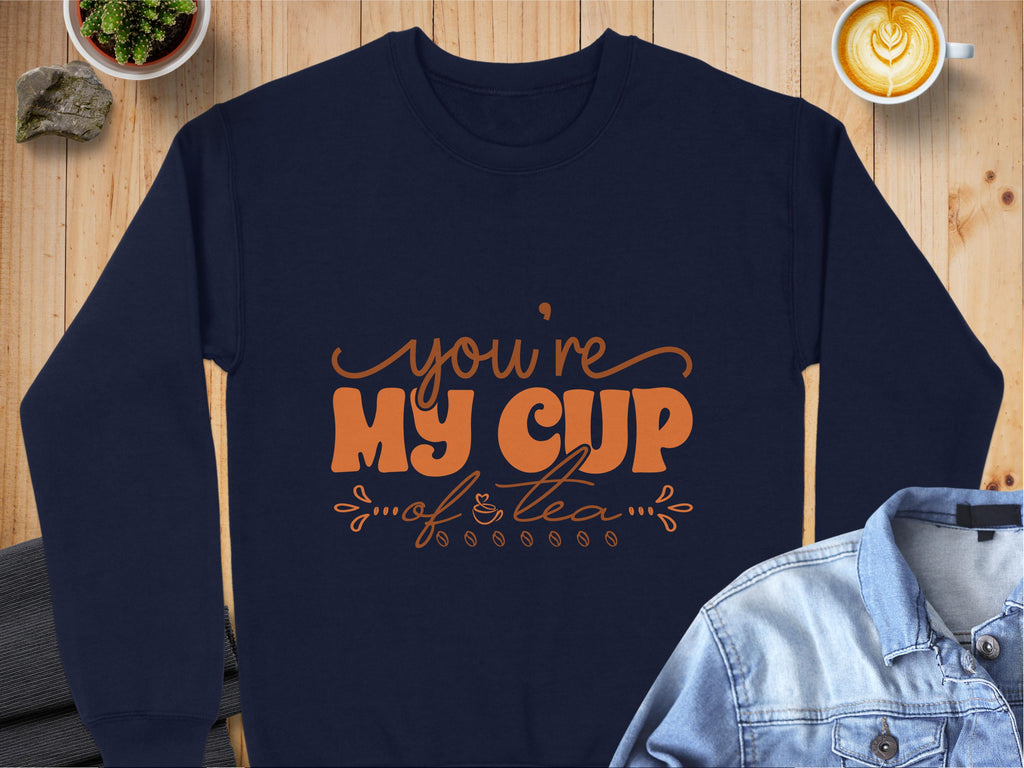 A navy sweatshirt by Garment Graphics features Youre My Cup of Tea in orange text, offering comfort with unisex sizing. Its displayed on a wooden table next to a denim jacket, heart-design latte, and small plant.