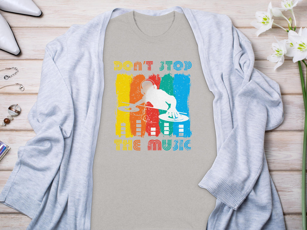 A Garment Graphics gray T-shirt with a vibrant DJ design and Dont Stop the Music text sits on a wooden surface. Ideal for music lovers, its accented by a light cardigan, white heels, jewelry, and white flowers.