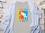 A Garment Graphics gray T-shirt with a vibrant DJ design and Dont Stop the Music text sits on a wooden surface. Ideal for music lovers, its accented by a light cardigan, white heels, jewelry, and white flowers.