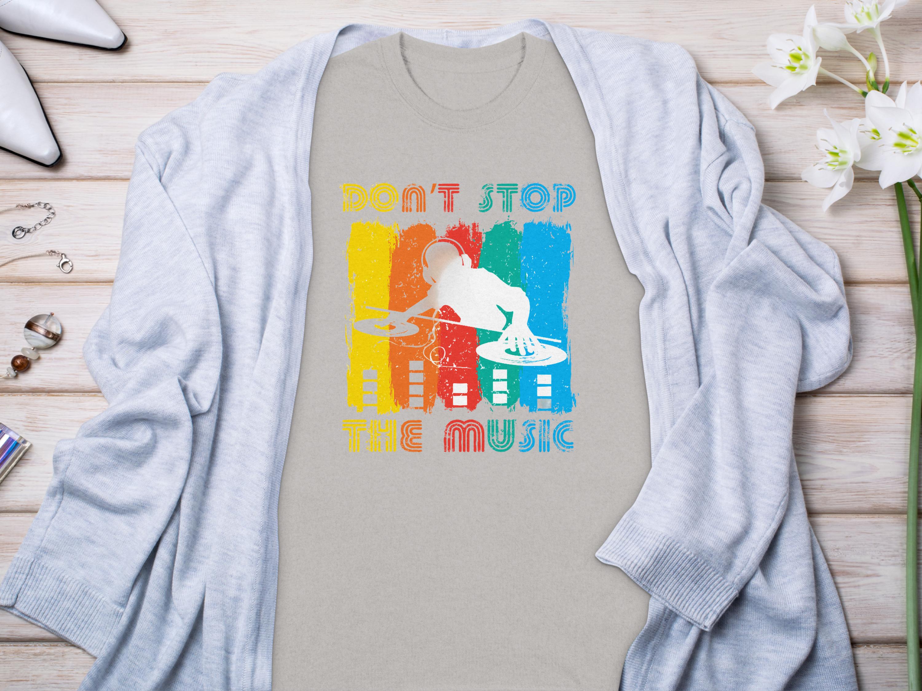 A Garment Graphics gray T-shirt with a vibrant DJ design and Dont Stop the Music text sits on a wooden surface. Ideal for music lovers, its accented by a light cardigan, white heels, jewelry, and white flowers.