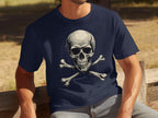 Wearing a navy blue graphic T-shirt with a striking skull and crossbones design by Garment Graphics, a person makes a bold fashion statement outdoors, sitting on a wooden bench. The dry grass and trees add a rustic backdrop to this edgy look.