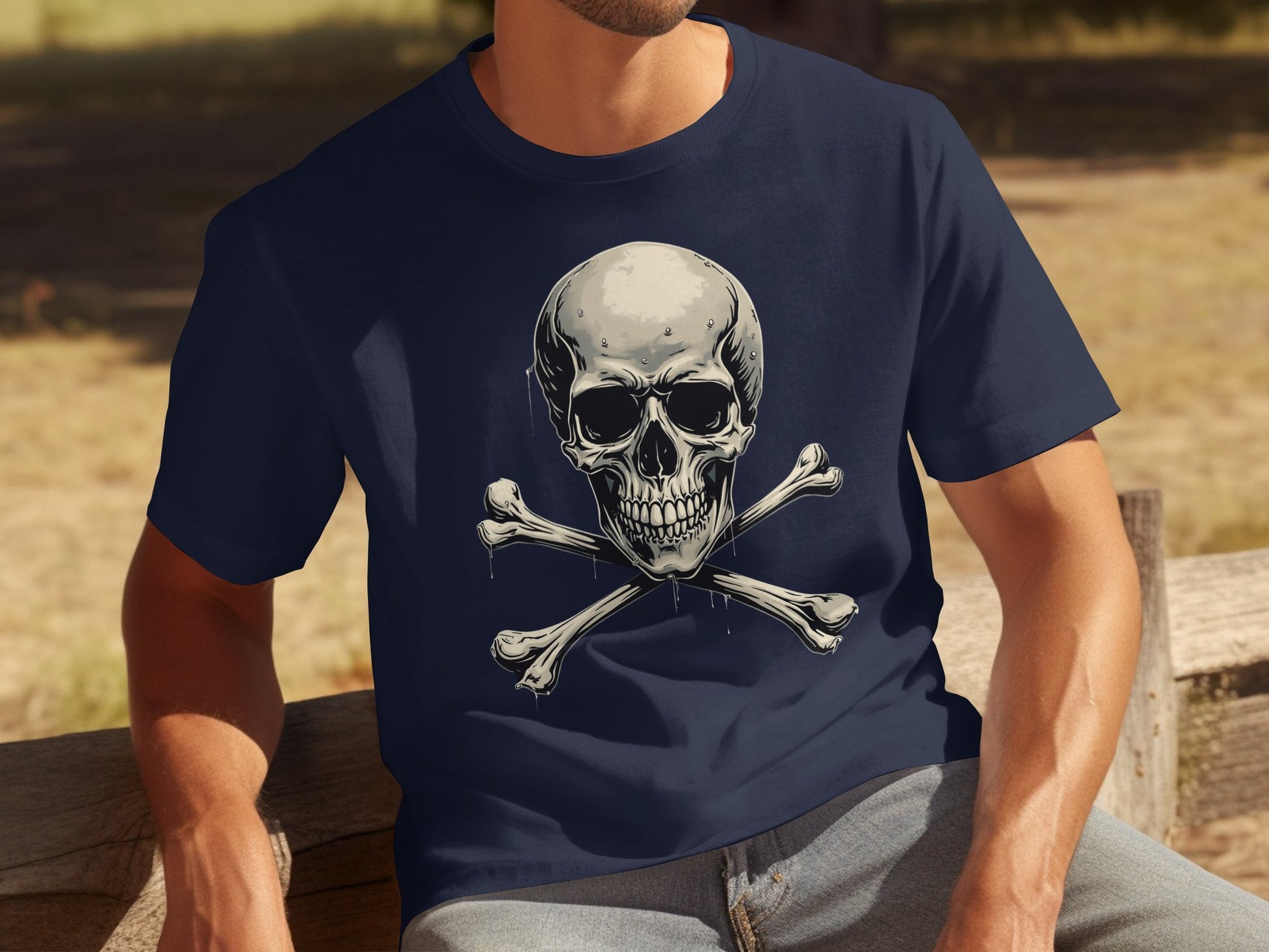 Wearing a navy blue graphic T-shirt with a striking skull and crossbones design by Garment Graphics, a person makes a bold fashion statement outdoors, sitting on a wooden bench. The dry grass and trees add a rustic backdrop to this edgy look.