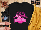 Flat lay of a Garment Graphics black sweatshirt with a pink Always Party Beach Girl design, showcasing three women and palm trees. Its surrounded by a gold jacket, makeup palette, sunglasses, shoes, perfume, and brushes on a wooden background.